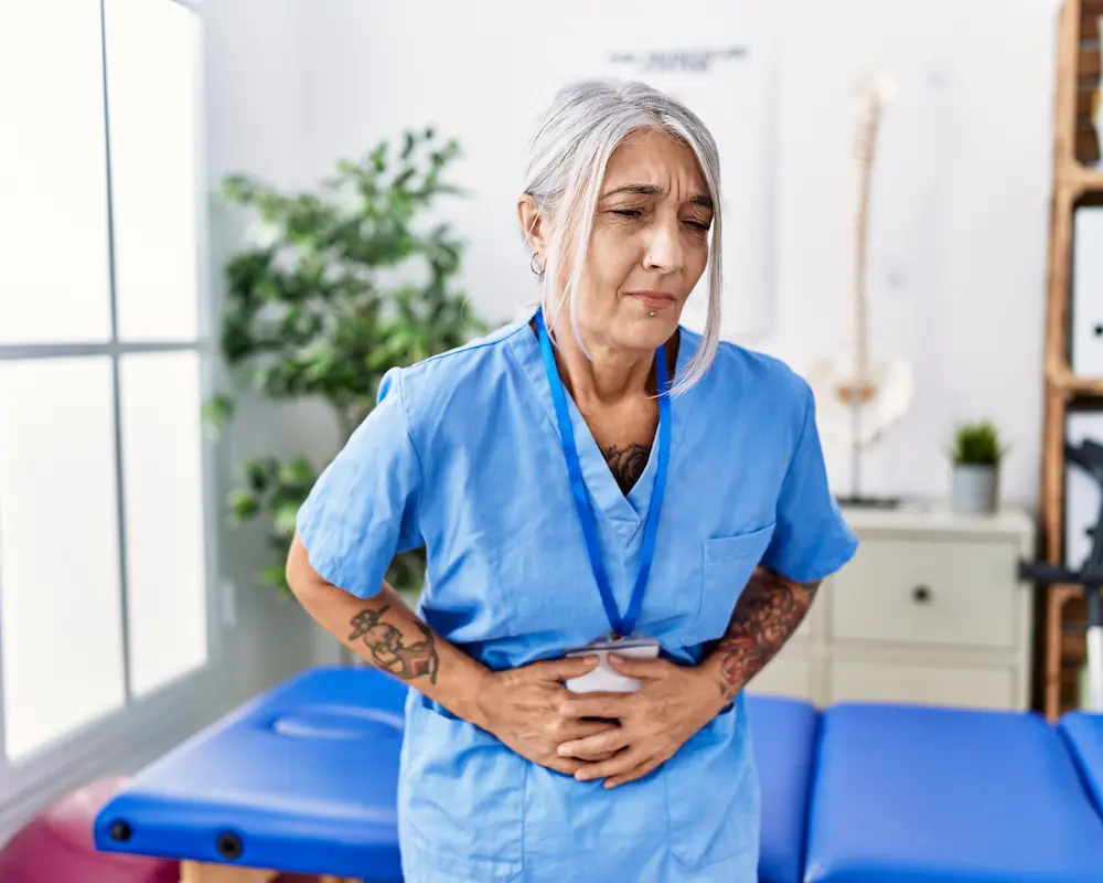 middle-age-greyhaired-woman-wearing-physiotherapist-uniform-medical-clinic-with-hand-stomach-because-nausea-painful-disease-feeling-unwell-ache-concept (1)